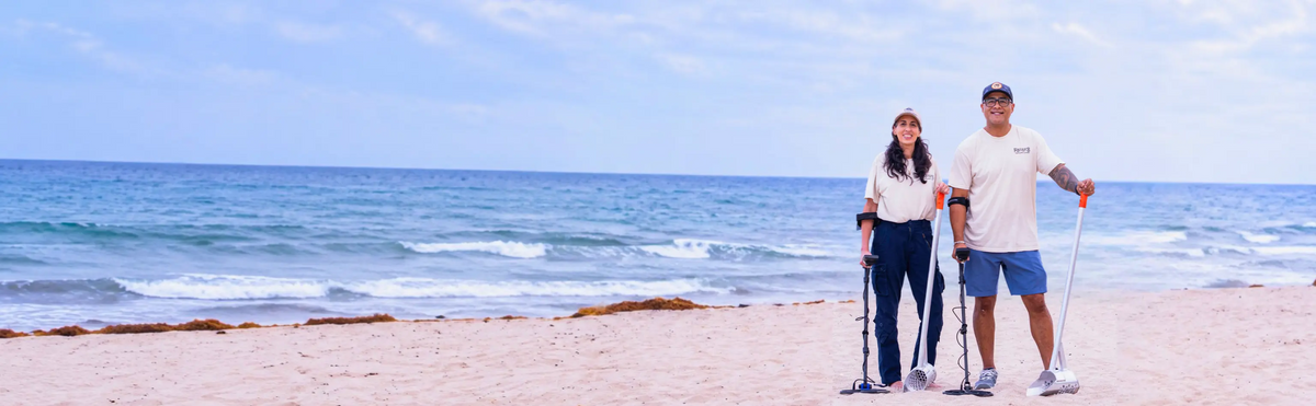 Two people metal detecting on a beach with sand scoops and the ocean in the background.