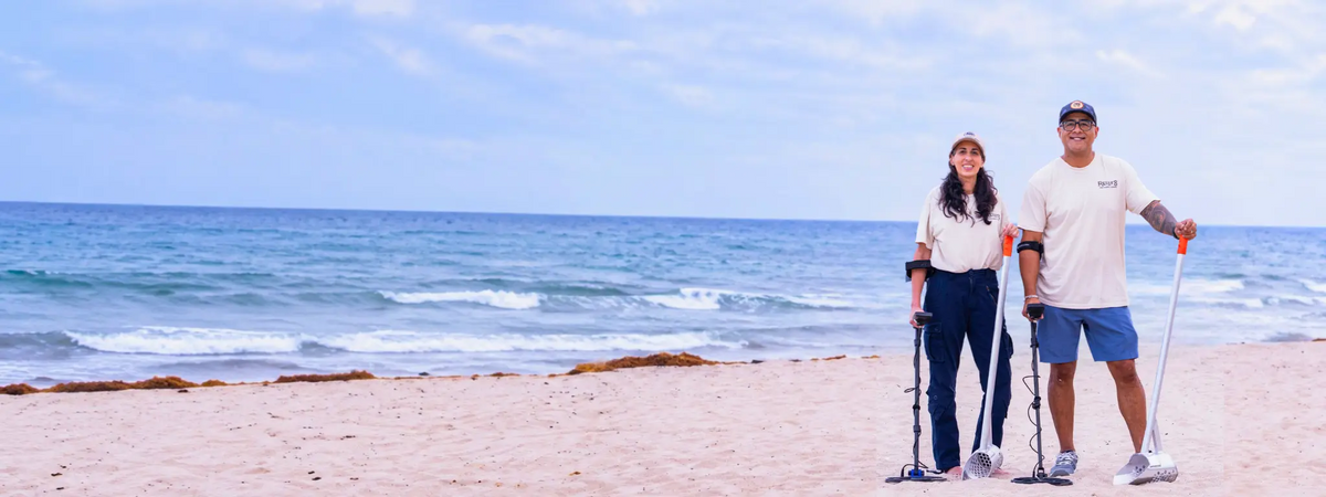 Two people metal detecting on a beach with sand scoops and the ocean in the background.